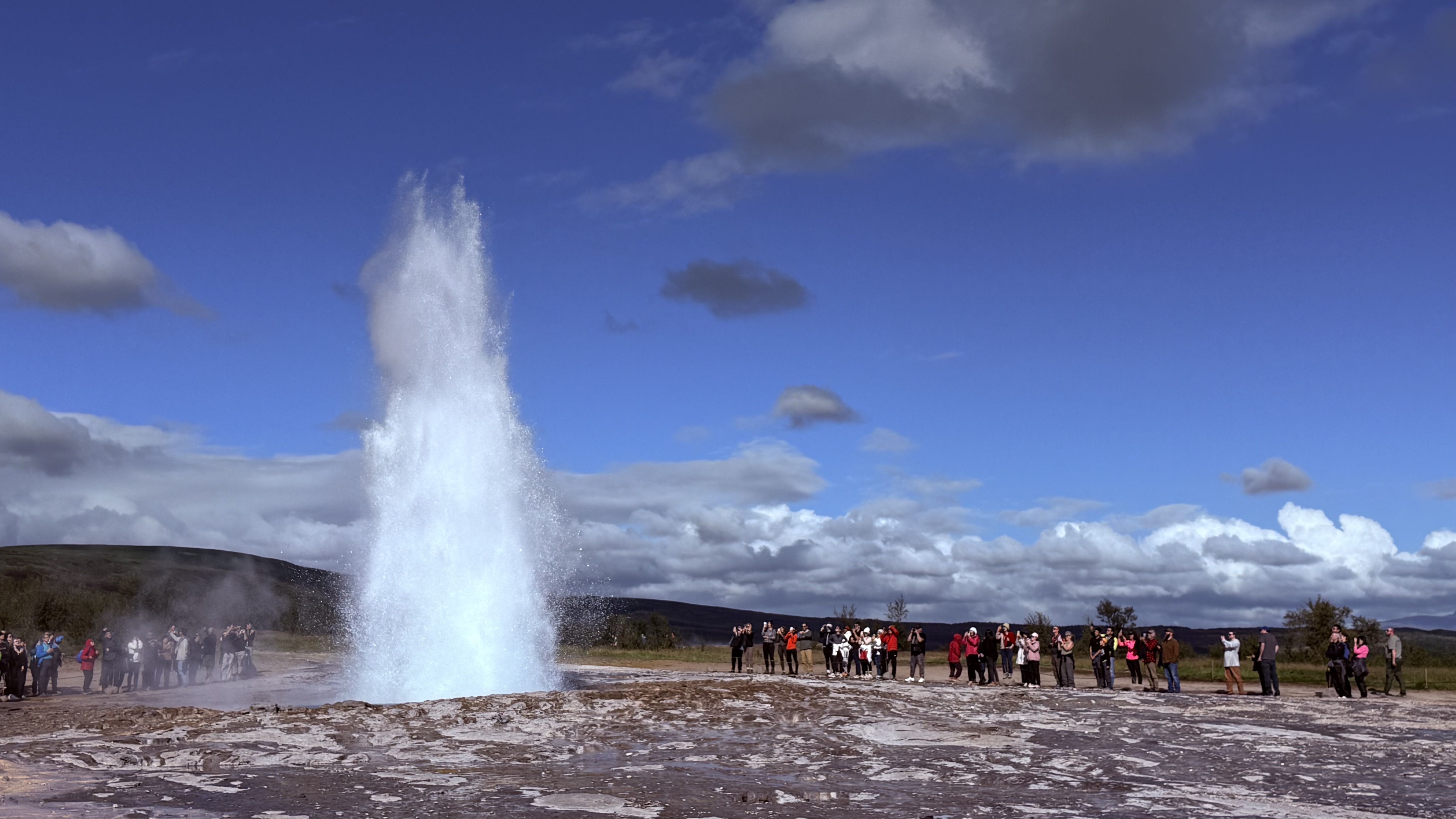 Strokkur geyser erupting against a bright blue sky with onlookers